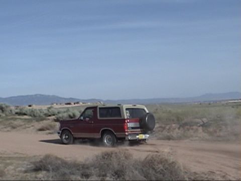 1994 Ford Bronco - 1994 Ford Bronco driving on a dirt road in a dry, open landscape under a clear blue sky.