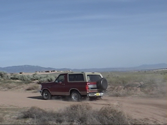 Maroon 1994 Ford Bronco with a tan top driving on a dirt road, kicking up dust. Spare tire mounted on the rear