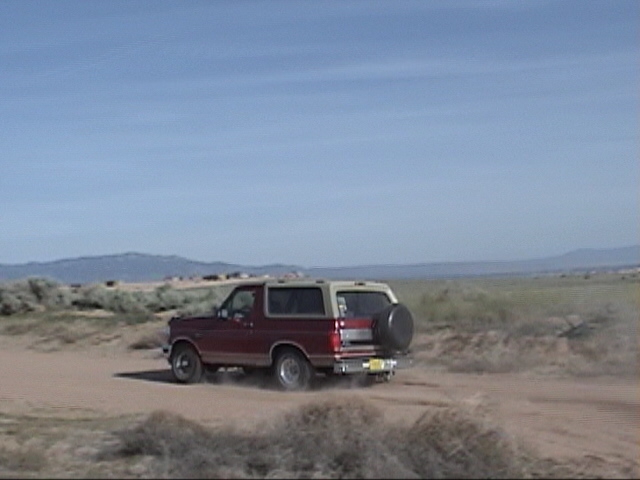 Red Ford Bronco with tan top driving on a dirt road, kicking up dust. Spare tire mounted on back