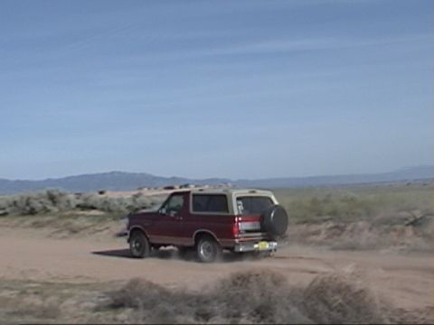 1994 Ford Bronco - A Ford Bronco kicks up dust as it drives across a dry, open landscape under a clear blue sky.