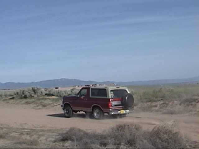 Red and tan 1994 Ford Bronco driving on dirt road, rear view
