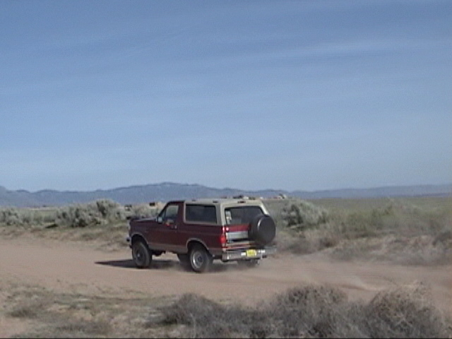 Red and tan Ford Bronco with a spare tire on the back, driving on a dirt road