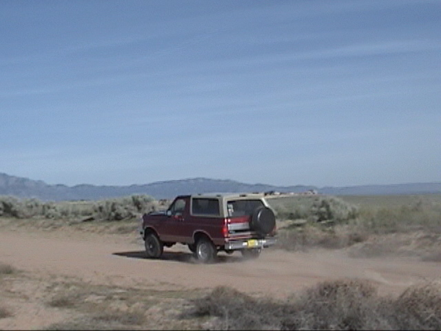 1994 Ford Bronco driving on a dirt road, kicking up dust. Spare tire mounted on the rear