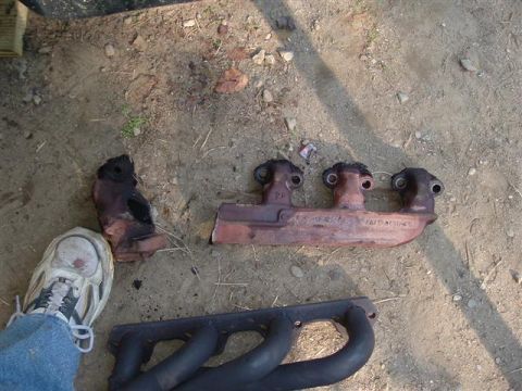 1991 Ford Bronco - Two exhaust manifolds, one rusty and broken, the other black, lie on the ground.