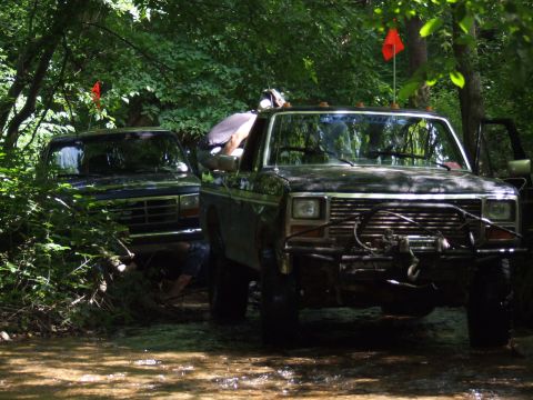 1986 Ford Bronco - Two SUVs navigate a wooded trail, one prominently a winch and custom bumper.