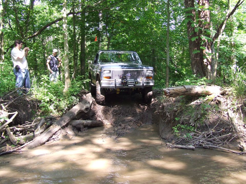 1986 Ford Bronco driving through muddy water and over logs in a wooded area