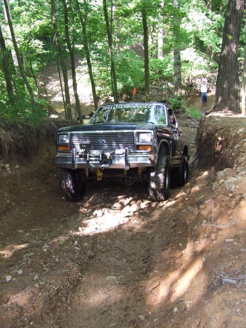 1986 Ford Bronco - A black Ford Bronco navigates a challenging off-road trail surrounded by trees.