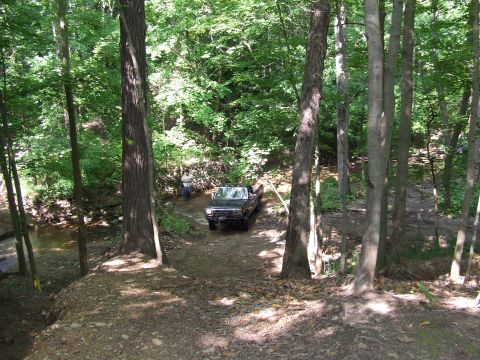 1986 Ford Bronco - A 1986 Ford Bronco navigates a wooded trail, with a person standing nearby.