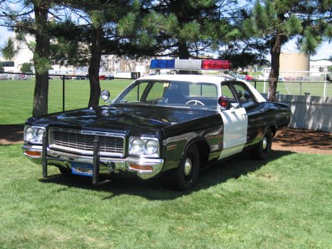 1991 Ford LTD Crown Victoria - black-and-white 1970s police sedan shown front three-quarter on grass, fitted with a chrome push bar and roof light bar.