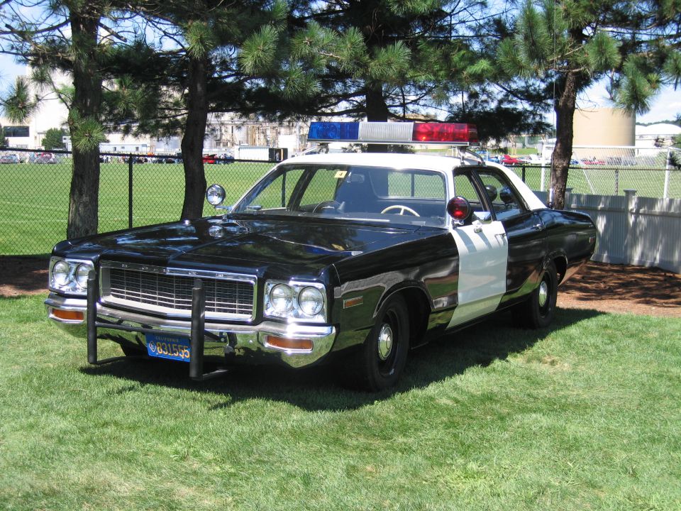 Front three-quarter view of a black and white 1970s police sedan with push bar and light bar