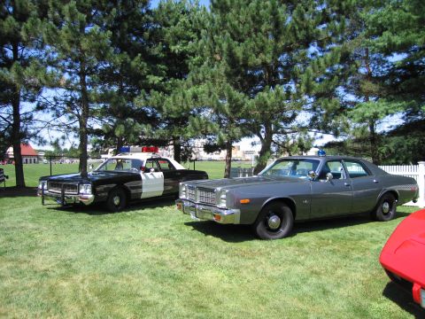 1991 Ford LTD Crown Victoria - black-and-white police sedan parked next to a gray full-size sedan on a grassy field, with a red car edge in the foreground.