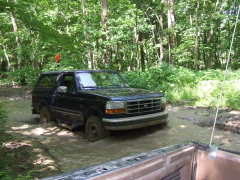 1986 Ford Bronco - A black Ford Bronco is shown driving through a muddy path in a wooded area.