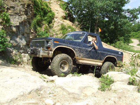 1986 Ford Bronco - A driver maneuvers a dark blue Ford Bronco through a challenging, rocky off-road course.