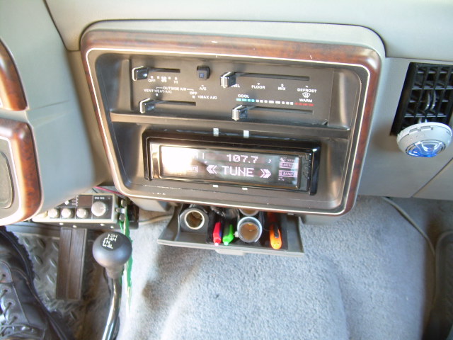 Car interior showing dashboard climate controls, an aftermarket radio tuned to 107.7, and a gear shift