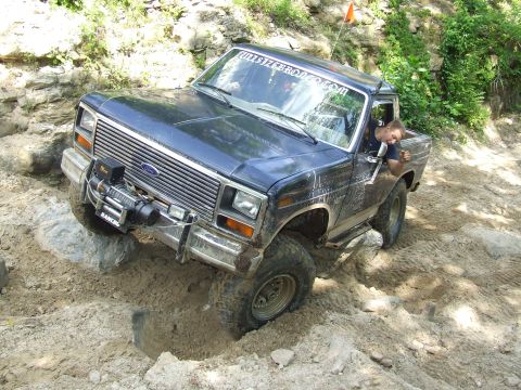 1986 Ford Bronco - A dark blue Ford Bronco navigates a challenging off-road course with a winch attached to its front bumper.