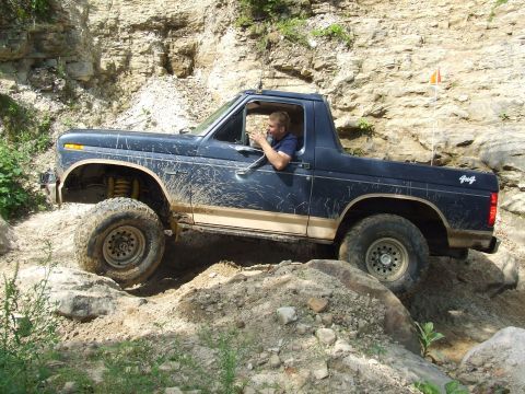 1986 Ford Bronco - Driver maneuvers a dark blue Ford Bronco through a challenging off-road course with rocky inclines.