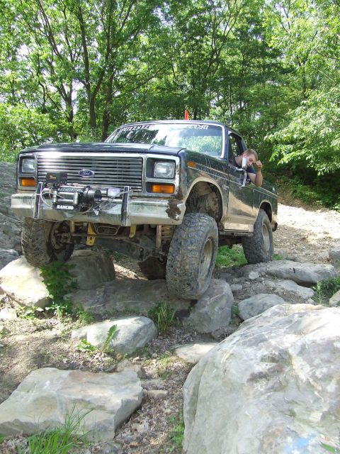 1986 Ford Bronco - A lifted Ford truck navigates a rocky trail with trees in the background.