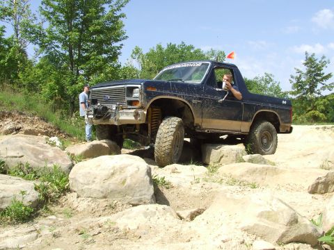 1986 Ford Bronco - A driver maneuvers a lifted Ford Bronco over large rocks on an off-road course.