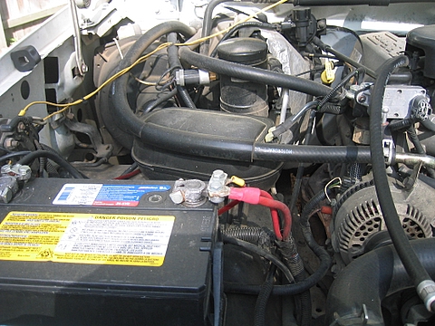 1996 Ford Bronco - Engine bay close-up of the driver-side battery with its yellow warning label, showing the clean lead terminals with a red-booted positive c…