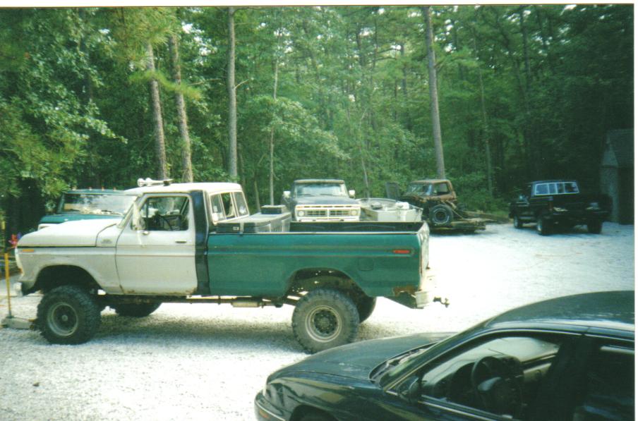 White and green lifted single-cab pickup truck parked on gravel, side view, surrounded by trees and other vehicles