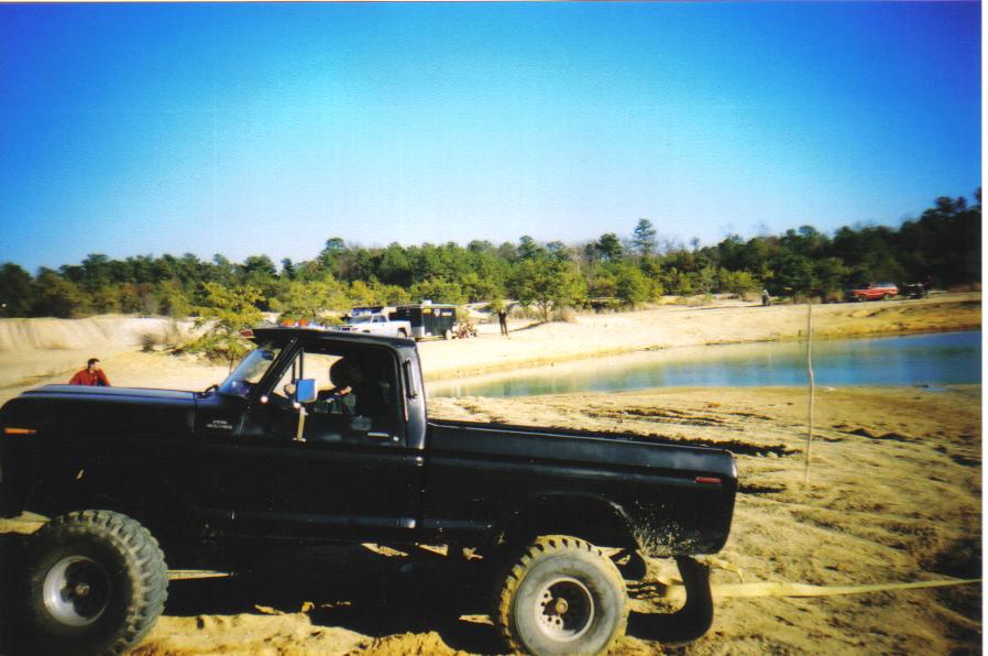 Black 1979 Ford F-250 pickup truck with large tires parked on sandy terrain near a body of water. Other vehicles and people are visible in the background