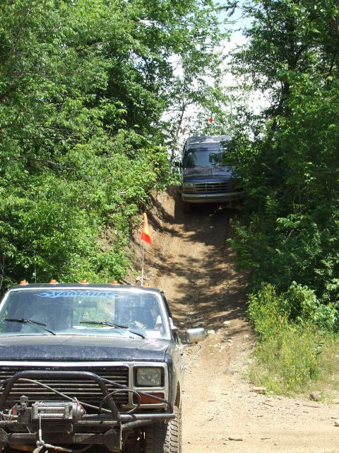 1986 Ford Bronco - Two off-road vehicles navigate a challenging dirt path through a wooded area on a sunny day.