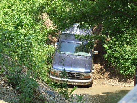 1986 Ford Bronco - A blue SUV is stuck in a muddy ditch surrounded by dense green foliage.