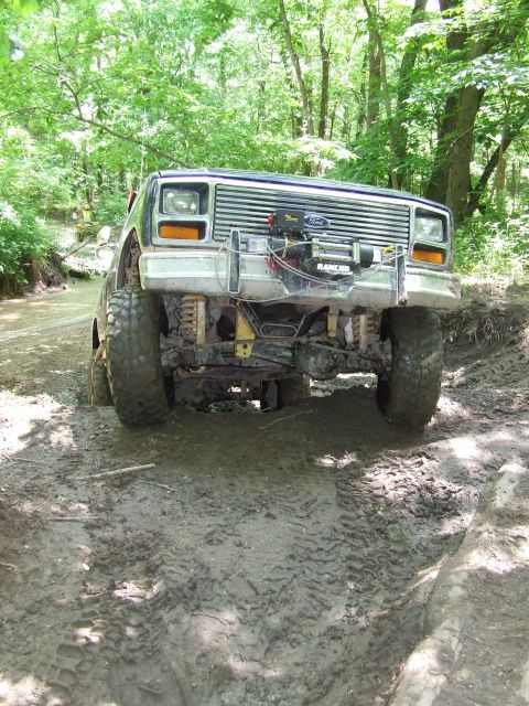 1986 Ford Bronco - A lifted Ford Bronco navigates a challenging muddy path in a wooded area.