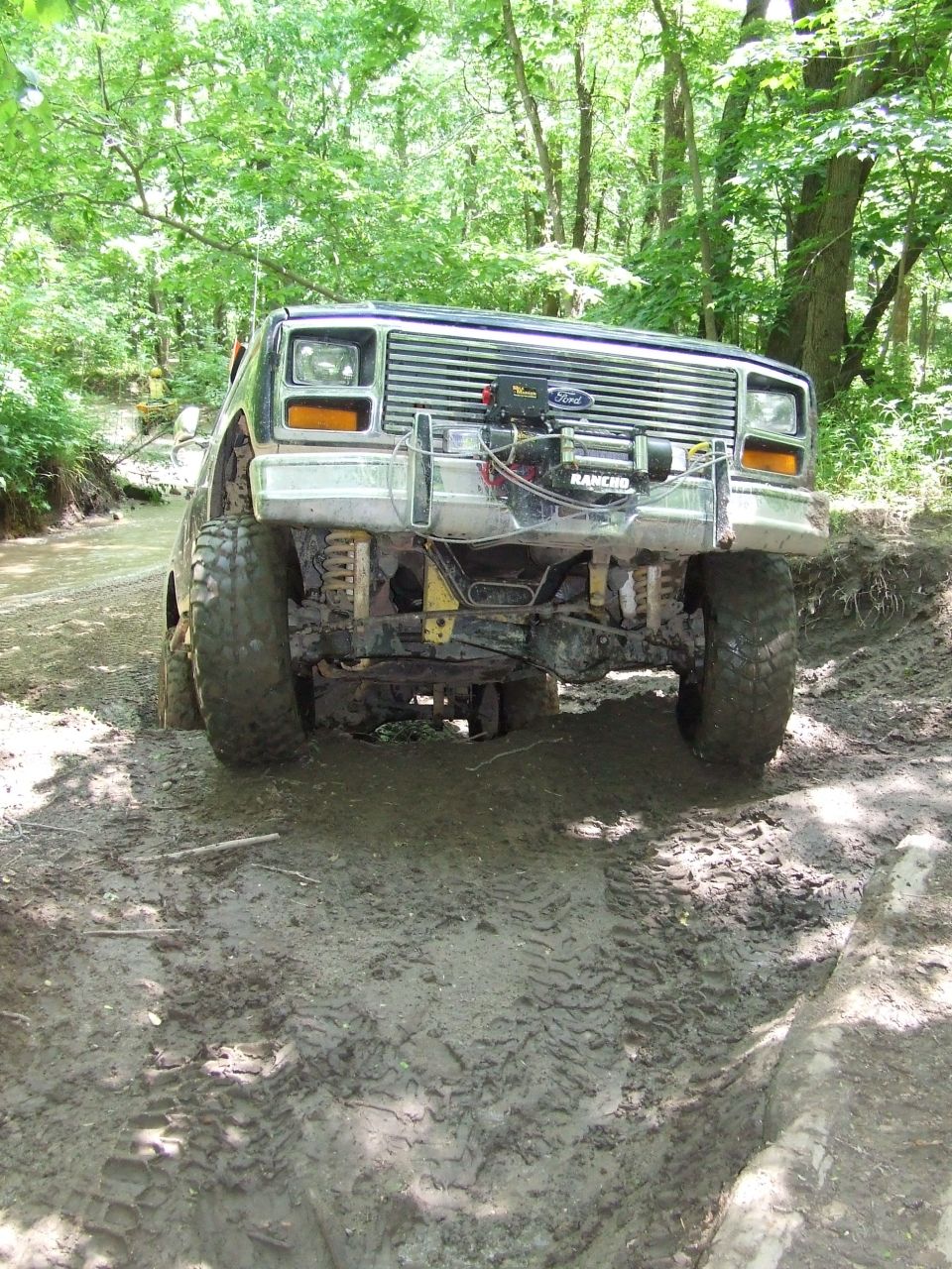 Mud-covered 1980s Ford Bronco with a winch on the front bumper, driving through a muddy forest trail