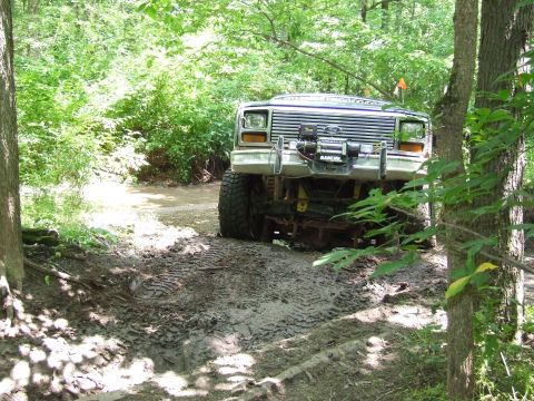 1986 Ford Bronco - Ford Bronco navigates a challenging muddy off-road course surrounded by dense trees.