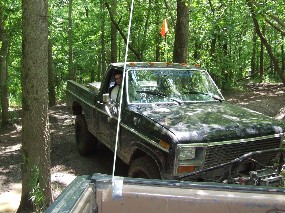 Black 1979 Ford F-150 pickup truck with winch, driving off-road in woods. Another vehicle's tailgate is in the foreground