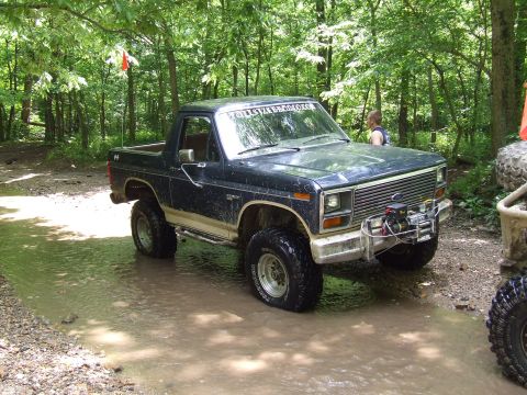 1986 Ford Bronco - This lifted Ford Bronco is navigating a muddy trail in a wooded area, with another off-road vehicle nearby.