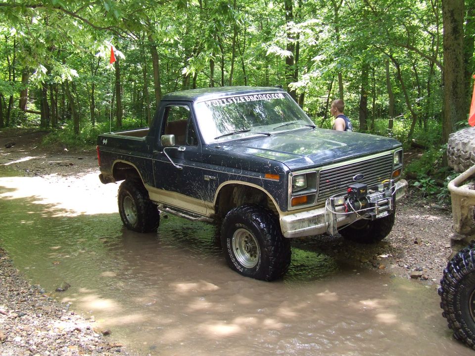 Dark blue Ford Bronco with a winch on front bumper driving through shallow water in woods. Another vehicle is partially visible on the right