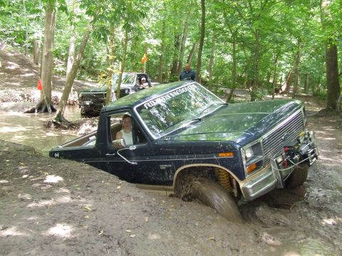 1986 Ford Bronco - A 1986 Ford Bronco navigates a muddy off-road course, with another vehicle visible in the background.