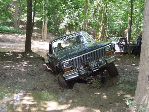 1986 Ford Bronco - A 1986 Ford Bronco navigates a muddy trail in a forest, with another vehicle visible in the background.