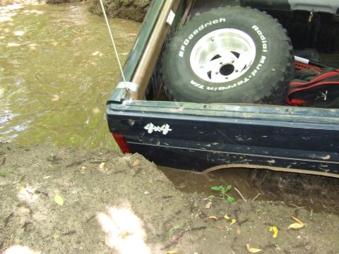 1986 Ford Bronco - A vehicle with a '4x4' decal is partially submerged in muddy water, with its spare tire visible in the bed.