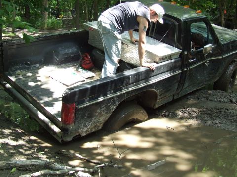 1986 Ford Bronco - A black pickup truck is stuck deep in the mud with a person reaching into the truck bed.