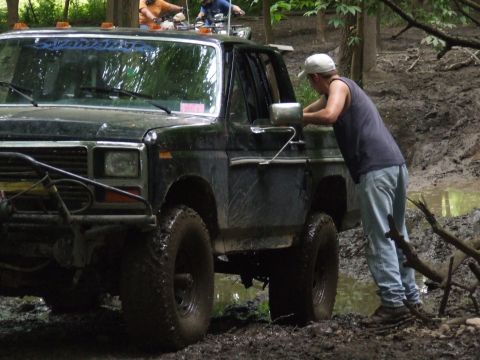 1986 Ford Bronco - A person checks on a muddy off-road vehicle while another vehicle is visible in the background.