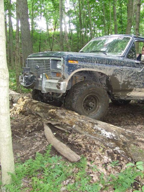 1986 Ford Bronco - A muddy Ford Bronco navigates a challenging off-road course through a wooded area.