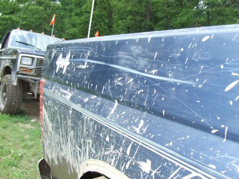 1986 Ford Bronco - Two muddy trucks are parked side-by-side in a grassy area with trees in the background.