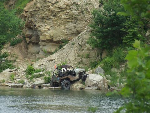 1986 Ford Bronco - Two off-road vehicles traverse a rocky shoreline next to a body of water, with a steep, layered rock formation in the background.