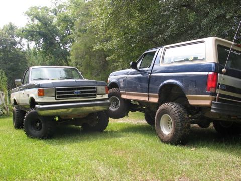 1989 Ford F-150 - Two lifted Ford trucks, a pickup and a Bronco, are parked side-by-side on a grassy area with trees in the background.