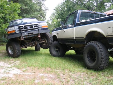 1989 Ford F-150 - Two lifted Ford trucks are parked on a grassy area with trees in the background.