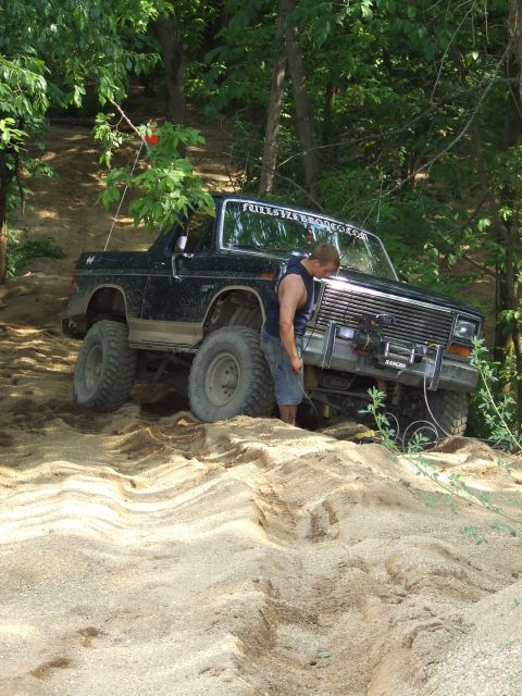 1986 Ford Bronco - A man inspects the front of a black Ford Bronco as it navigates a sandy, wooded trail.