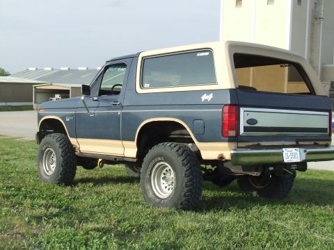 1986 Ford Bronco - Driver-side 3/4 rear view of a lifted two-tone dark blue and tan full-size Ford Bronco parked on grass beside a metal farm building, with a…