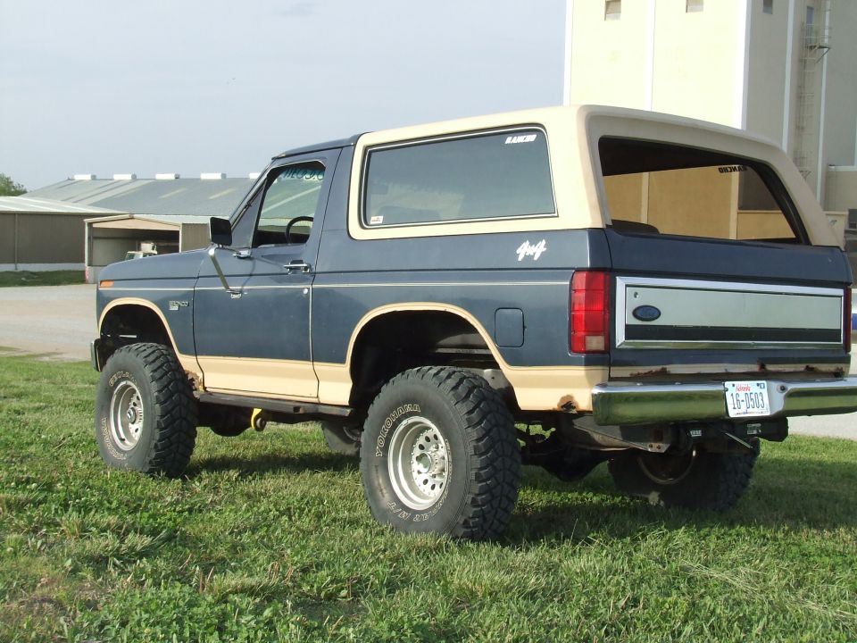 Lifted blue and tan Ford Bronco 4x4, rear 3/4 view on grass with white wheels and mud tires