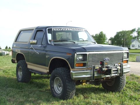 1986 Ford Bronco - Three-quarter front view of a lifted dark blue and tan two-tone square-body Ford Bronco parked on grass, wearing Ground Hawg mud-terrain ti…