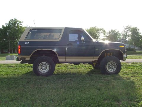 1986 Ford Bronco - Full side profile of a lifted dark blue and tan two-tone square-body Ford Bronco standing on grass, with a tan removable hardtop carrying a…