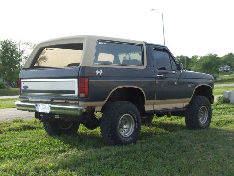 1986 Ford Bronco - Rear three-quarter view of a lifted two-tone dark blue and tan full-size Ford Bronco parked on grass, wearing a tan removable hardtop with…