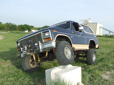 1986 Ford Bronco - Lifted two-tone blue and tan full-size Ford Bronco flexing with its passenger front tire climbing a concrete block in a grassy field, showi…